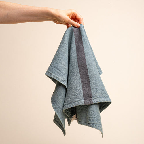 A hand holds the center of a linen towel in a blue-grey color with a dark grey stripe in the middle