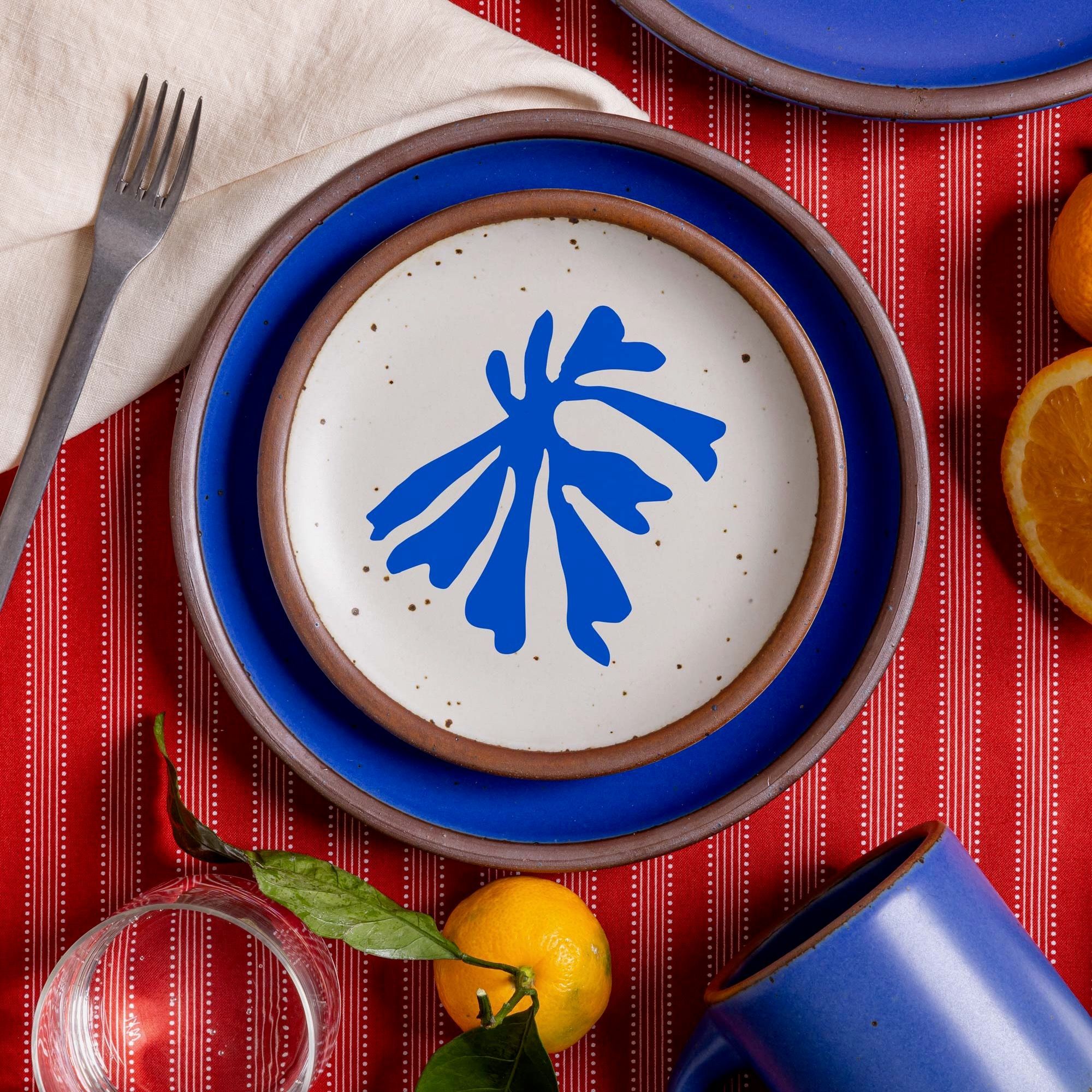 A stack of plates including one that has a blue frond-like cut-out layered with a ceramic plate in a cobalt blue color on a red tablecloth with oranges, a fork, and glass.