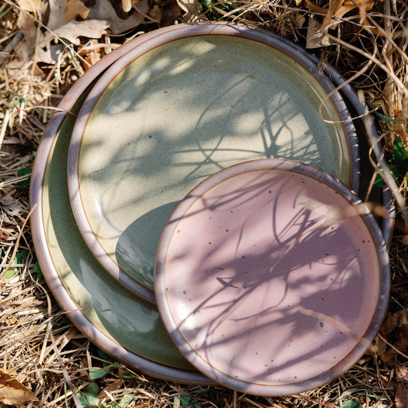 3 Plates of varying sizes in reflective dappled grey-green and dusty mauve pink artfully arranged in dried grass with shadows.