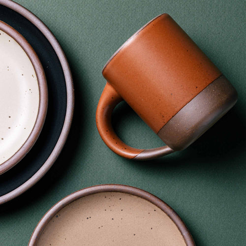 A medium sized ceramic mug with handle in a cool burnt terracotta color, surrounded by plates in various sizes.