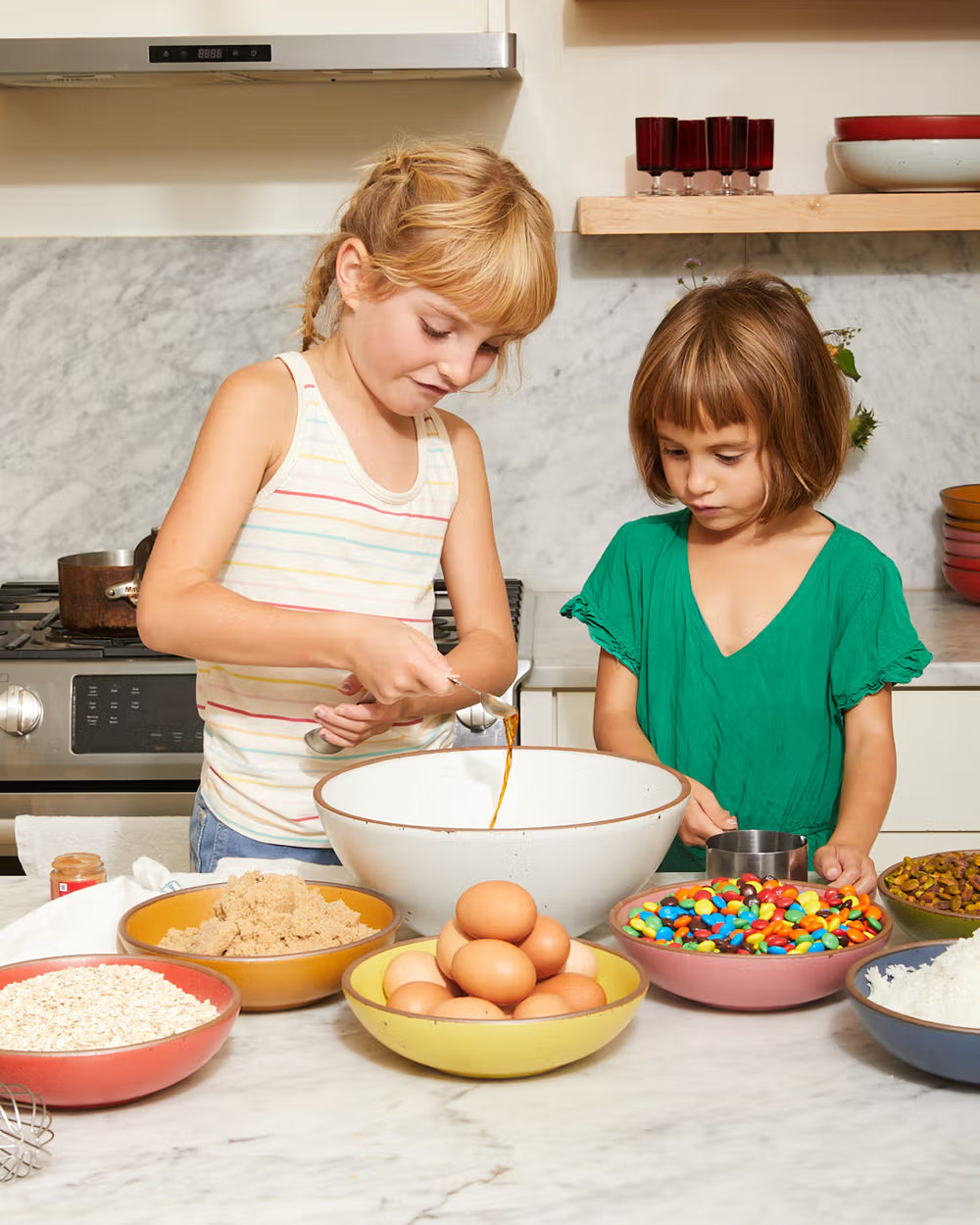 Two children are baking in a kitchen, surrounded by bowls of ingredients including eggs, brown sugar, M&M's, oats, and nuts on a marble countertop.