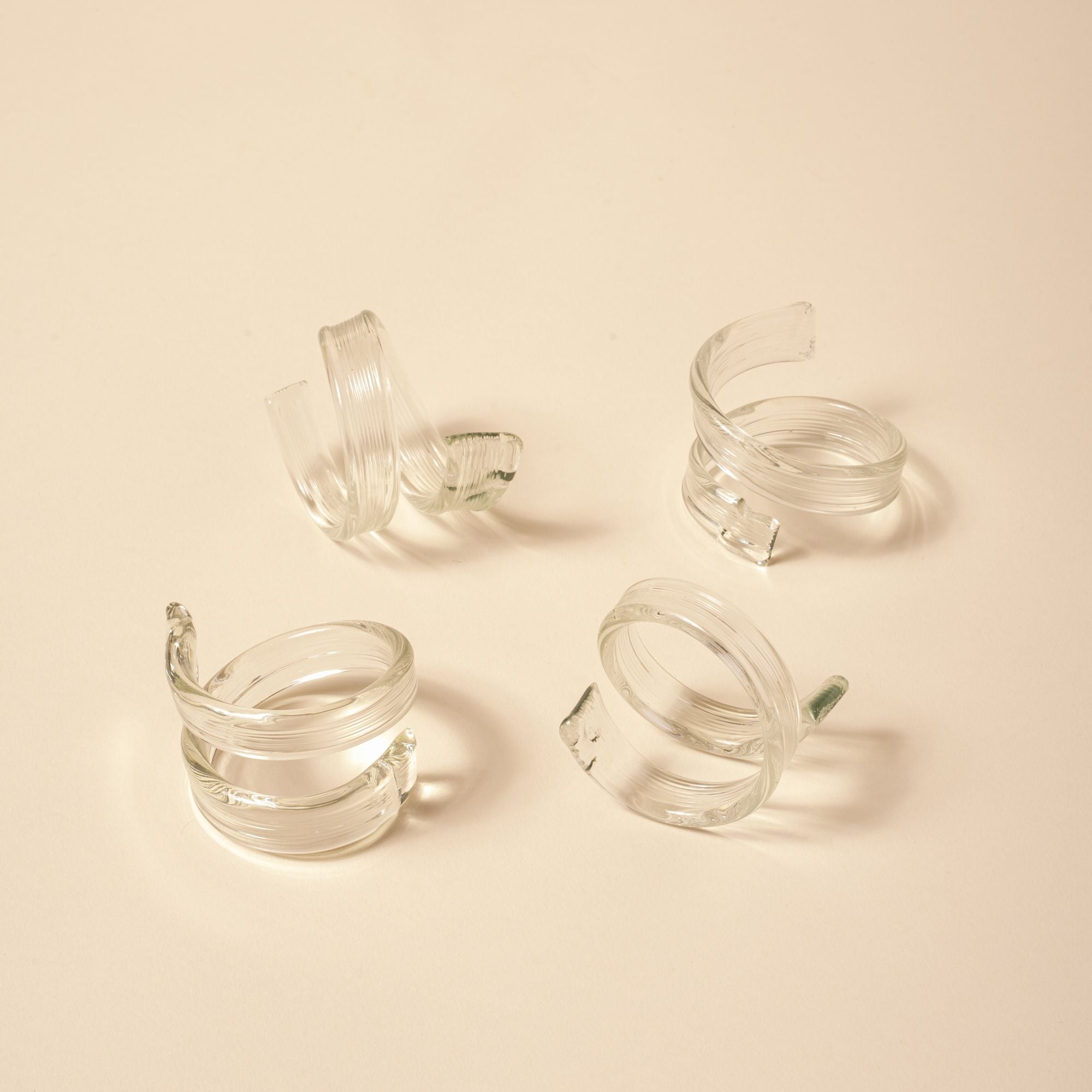 Set of four clear glass napkin rings with a coiled, ribbon-like design on a neutral background.
