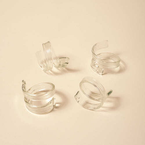 Set of four clear glass napkin rings with a coiled, ribbon-like design on a neutral background.