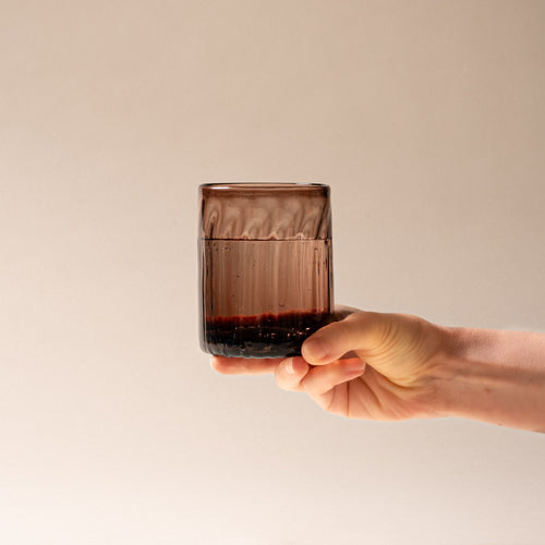 A hand holds out a taller amber-brown glass tumbler with vertical ridges, filled with water, set against a neutral background.