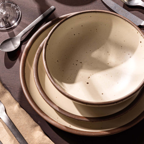 Close up of a table setting featuring a ceramic dinner plate, side plate, and bowl stacked  in a warm, tan-toned, off-white color