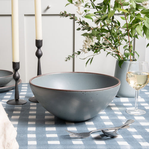 A large rounded ceramic bowl in a blue-grey color on a crosshatch tablecloth with a glass of wine.