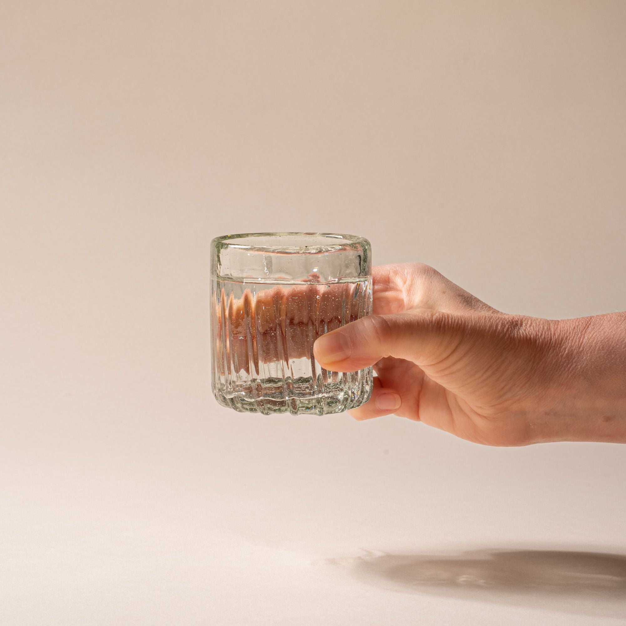 A hand holds out a clear textured glass tumbler with vertical ridges, filled with water, against a neutral background.