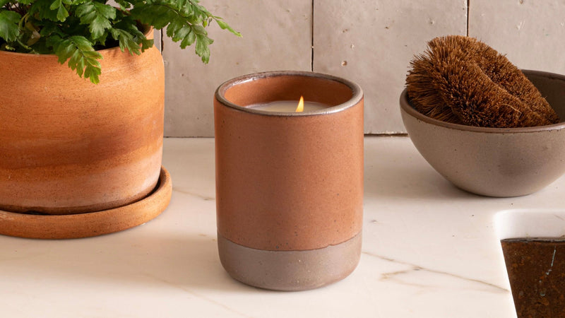 A lit candle in a brown ceramic holder is placed on a kitchen countertop with a terracotta plant pot and a bowl holding a scrub brush nearby.