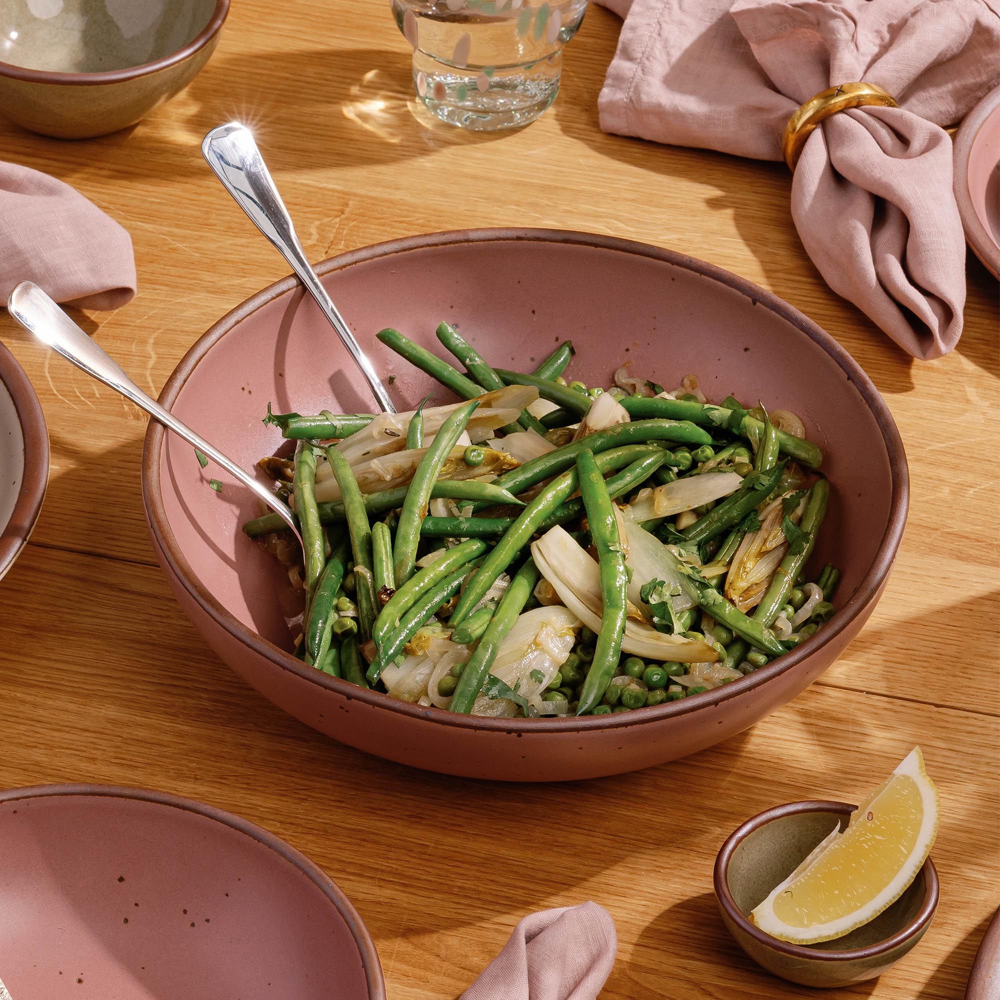 Large dusty mauve pink ceramic serving bowl filled with green bean salad, set on a wooden table with utensils and linen napkins.