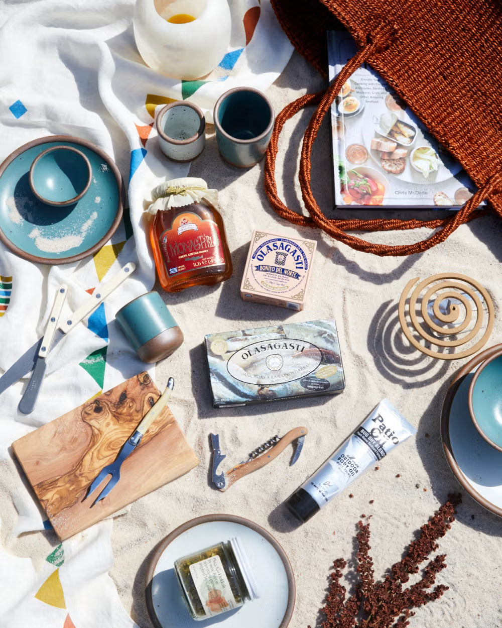 A beach scene with various items arranged on a sandy surface: ceramic plates and cups with a turquoise glaze, a wooden cutting board with a cheese fork, a jar of honey, tins of fish, a corkscrew, a tube of body cream, and a jar of pickles. A colorful patterned blanket, wicker tote, candle holder, and magazine are also visible.