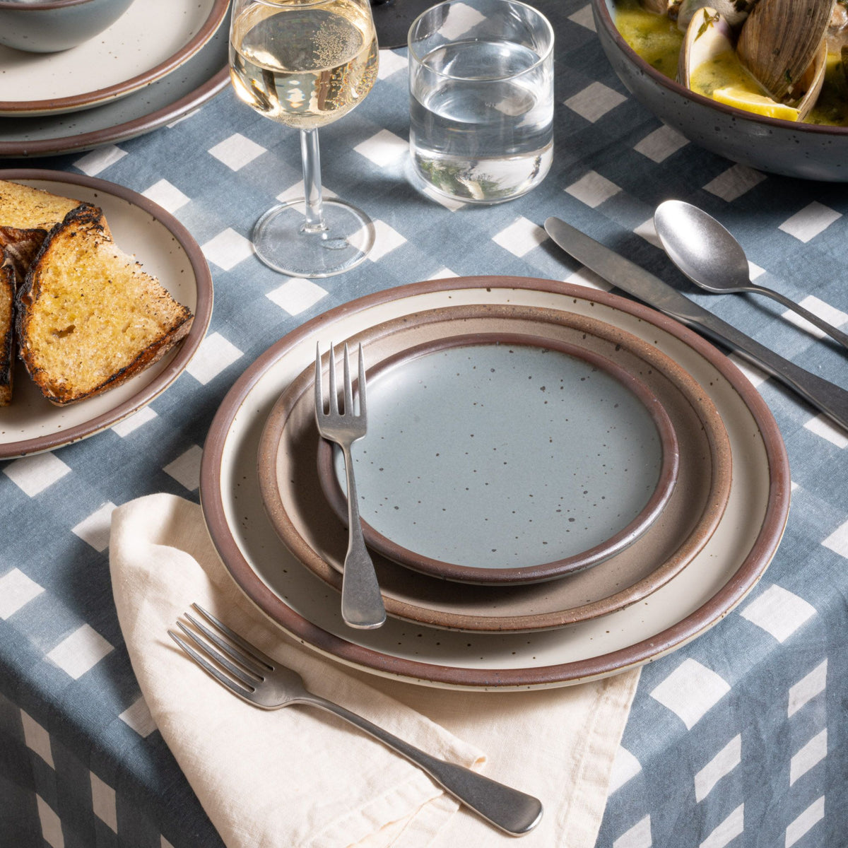 A place setting with ceramic dinnerware, tumbled matte steel flatware, on a blue-grey crosshatch tablecloth