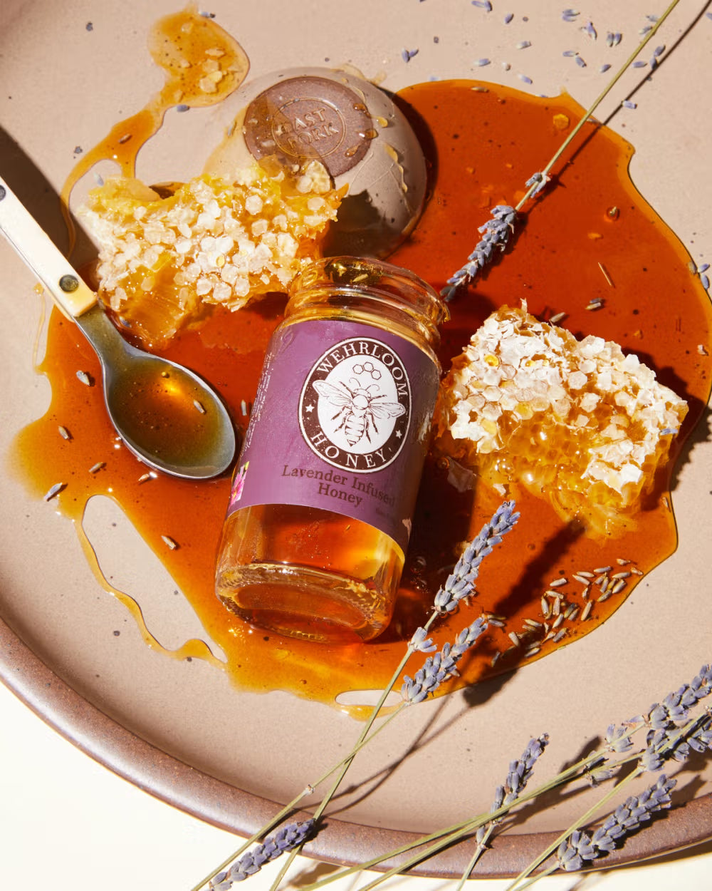 Jar of lavender-infused honey, honeycomb pieces, and a spoon with honey on a beige plate. Lavender sprigs are scattered around with honey drizzled across the surface.