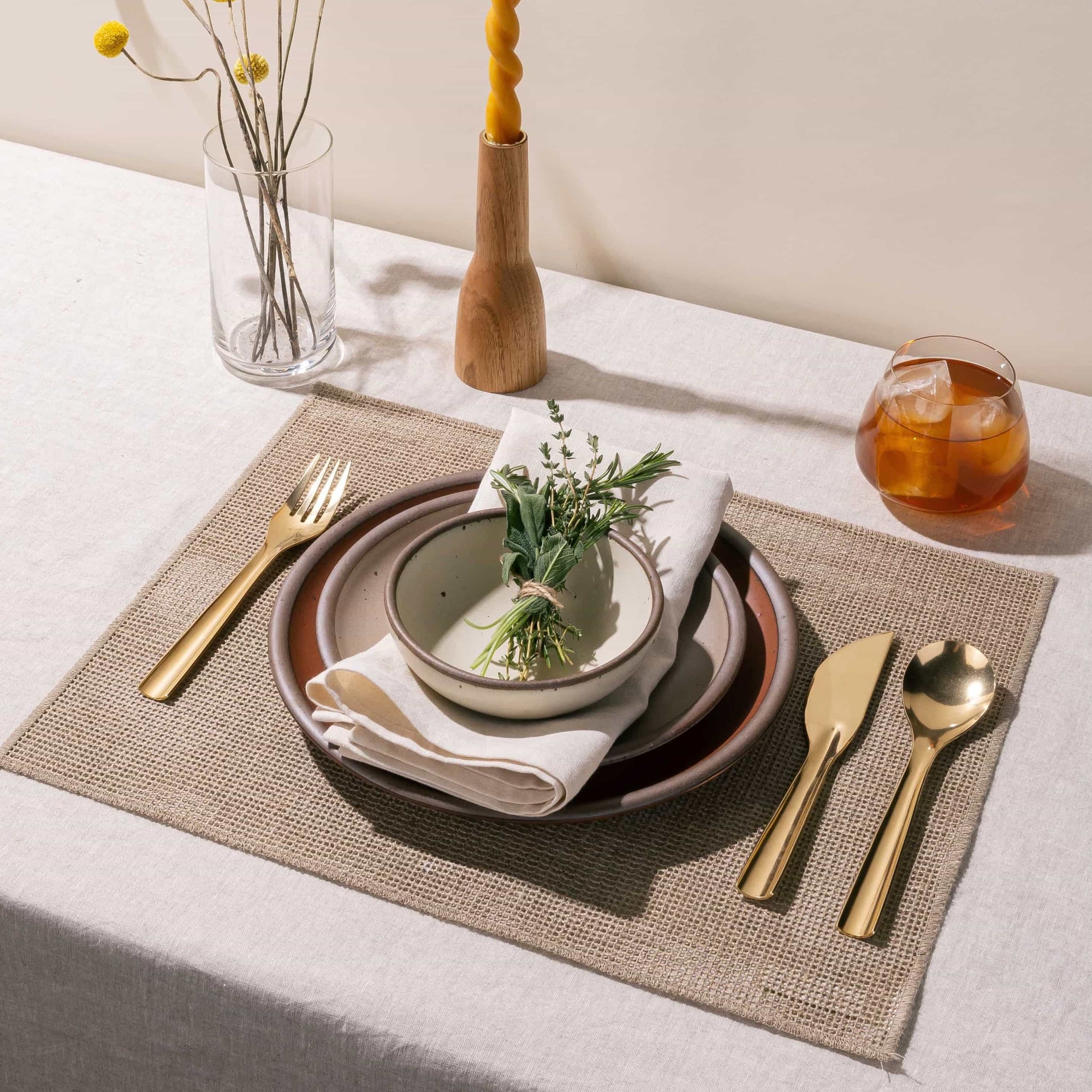 A table setting featuring a stack of ceramic plates, brass flatware, drink, taper candle, and more.