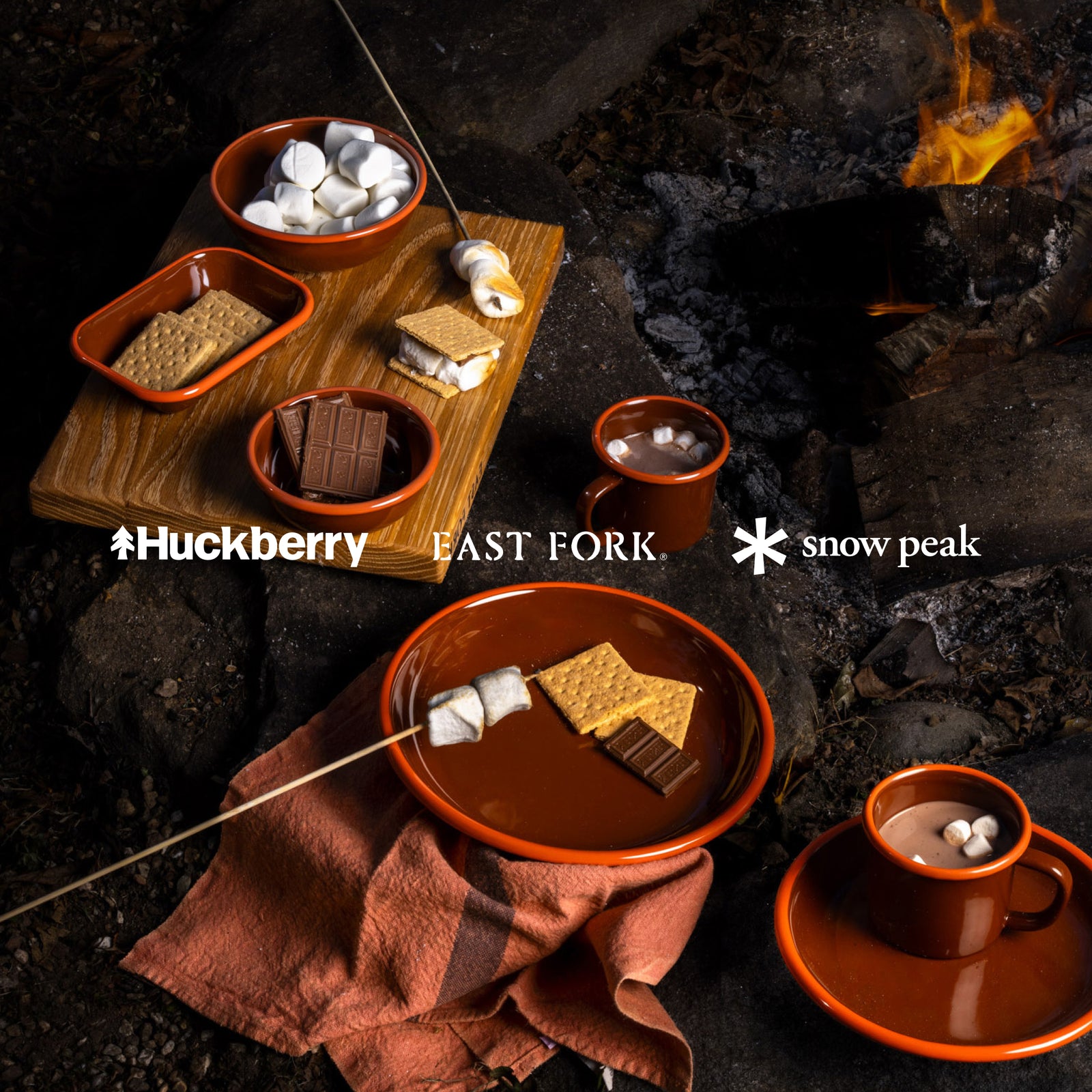 Colorful red enamelware plates, bowls, dishes and mugs around a campfire making s'mores, with overlay text that reads 'Huckberry, East Fork, Snow Peak'