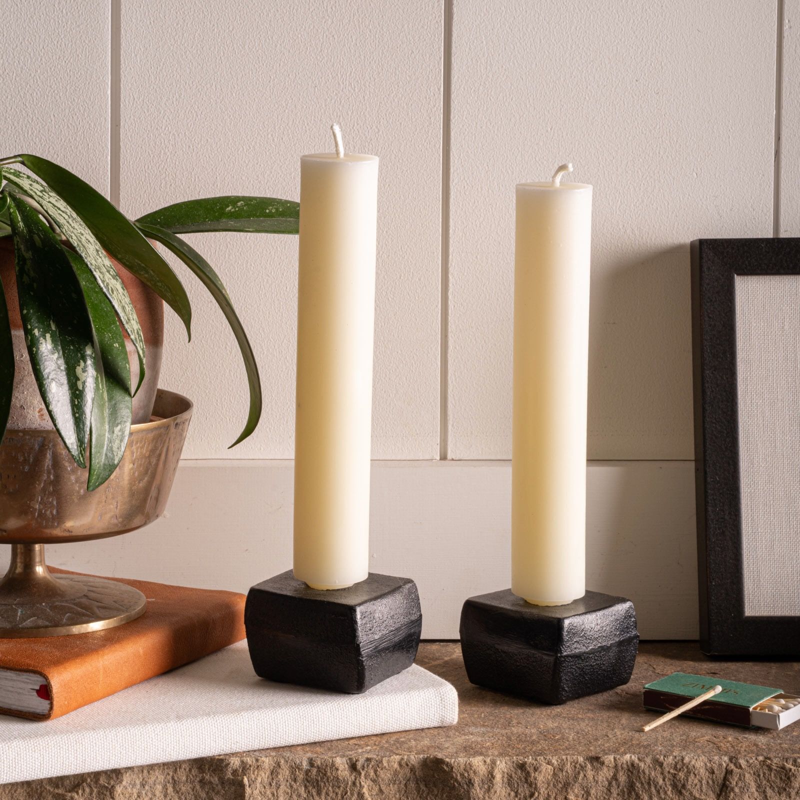 Two cream-colored column candles in black geometric holders sit on a stone ledge beside books, a potted plant, and a box of matches.