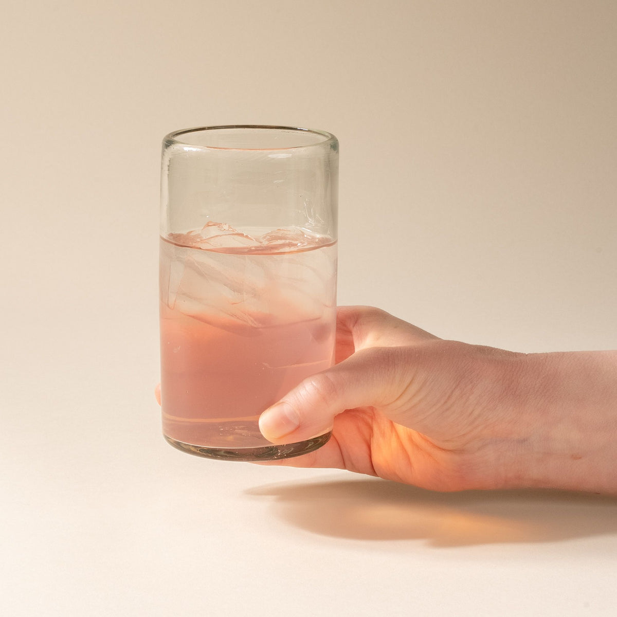 A hand holds a tall clear drinking glass, filled with pink lemonade.