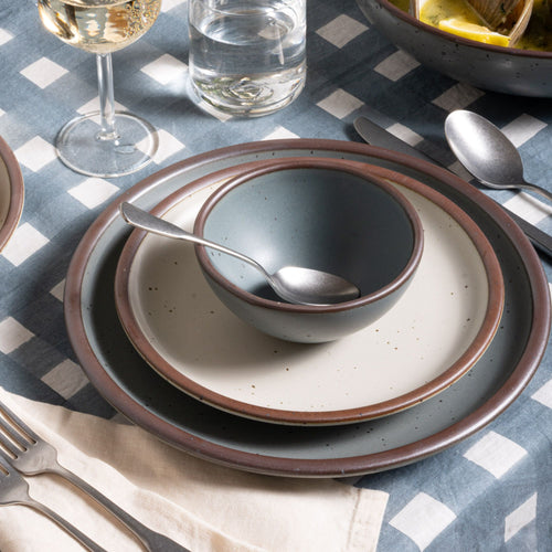 Small round bowl and dinner plate in a blue-grey color with a cream side plate, with silverware on a blue-grey crosshatch tablecloth