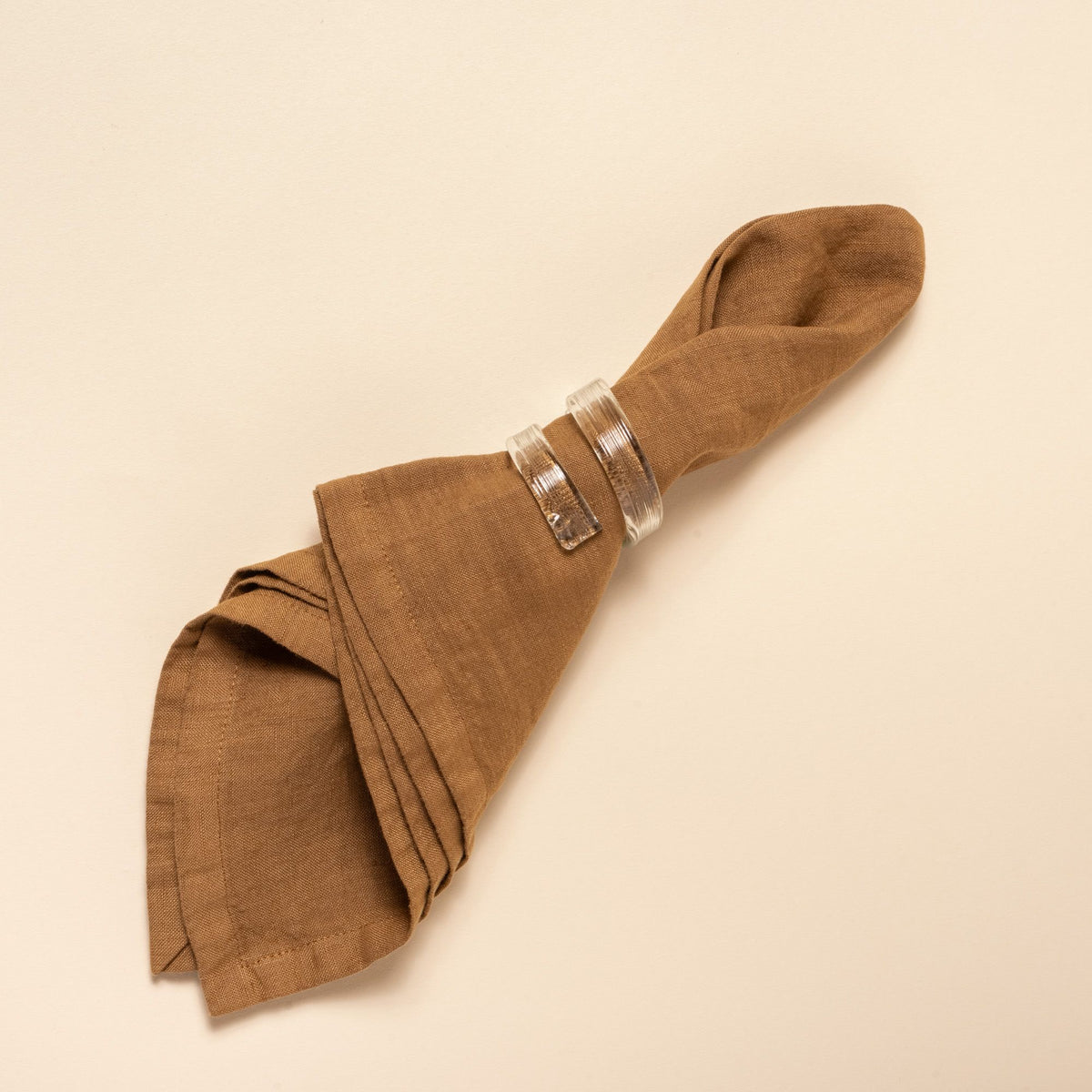 Brown linen napkin folded with curved glass clear napkin rings, styled against a neutral background.