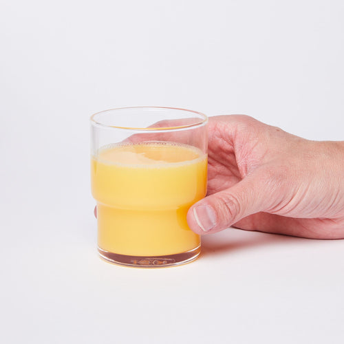 Hand holding a clear cylindrical glass with a wider top half with a narrower bottom half, full of orange juice