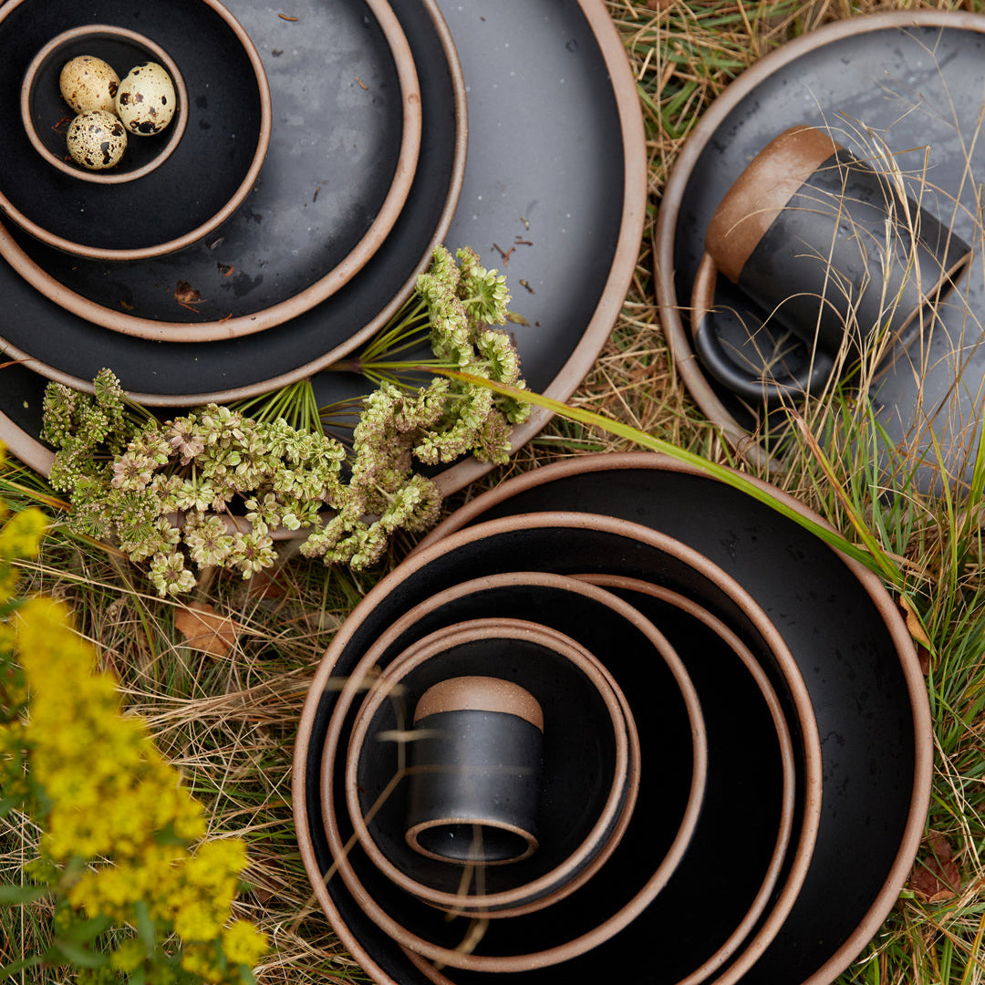 An overhead view of ceramic plates, bowls, and cups in various sizes with unglazed rims and glazed in a graphite black color.