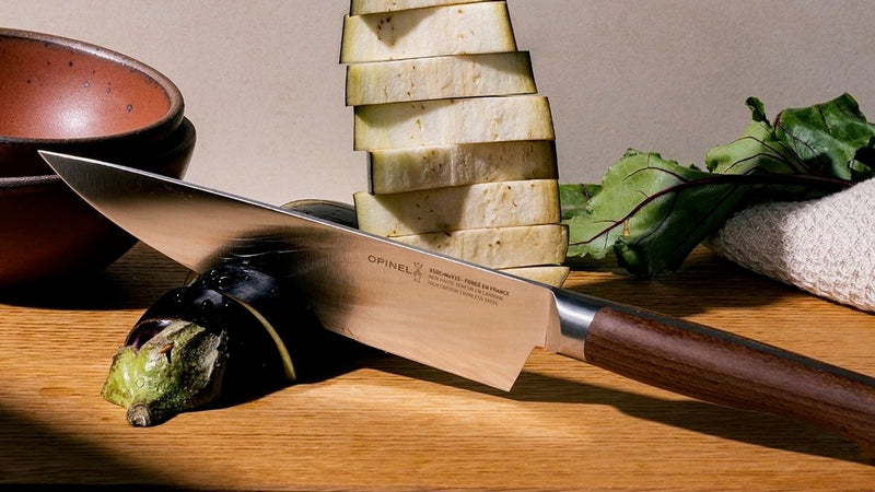 A chef's knife is propped up on a cutting board with slices of eggplant next to the blade and towered behind. The knife has a sharp steel blade with a beveled dark wood handle