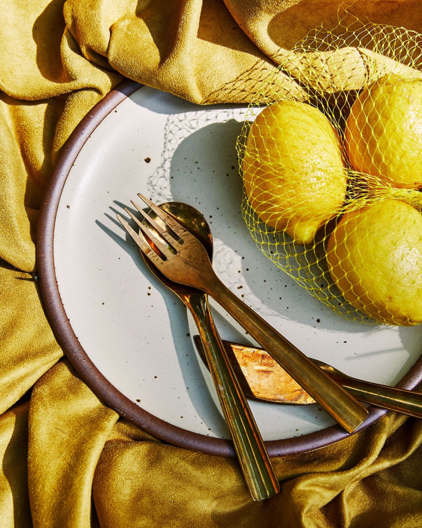 A shiny brass fork, knife and spoon lay on a ceramic off-white plate with a net of lemons and a gold fabric background