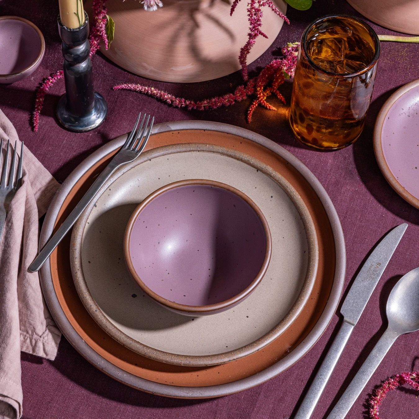 A place setting of terracotta, pale light brown, and light purple with silverware and a tortoiseshell tumbler glass