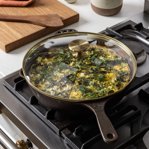 On a stove, there's a glass skillet lid with a brass lip and brass handle knob over a cast iron skillet. The skillet is filled with a casserole.
