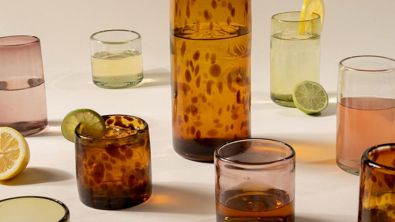 Amber speckled glass pitcher surrounded by colorful drinking glasses, citrus slices, and cocktail garnishes on a white surface.