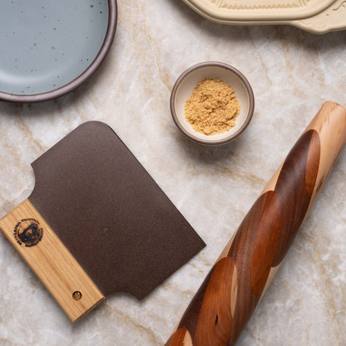 A marble surface displays a wooden rolling pin, a dough scraper with a wooden handle, a small bowl of brown sugar, and ceramic plates in neutral tones.