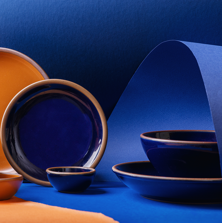 A styled studio arrangement of ceramic tableware in two contrasting glazes: a dark glossy blue and a warm earthy orange. The set includes plates and bowls, some nested together and others displayed upright, set against a split blue and orange paper background.