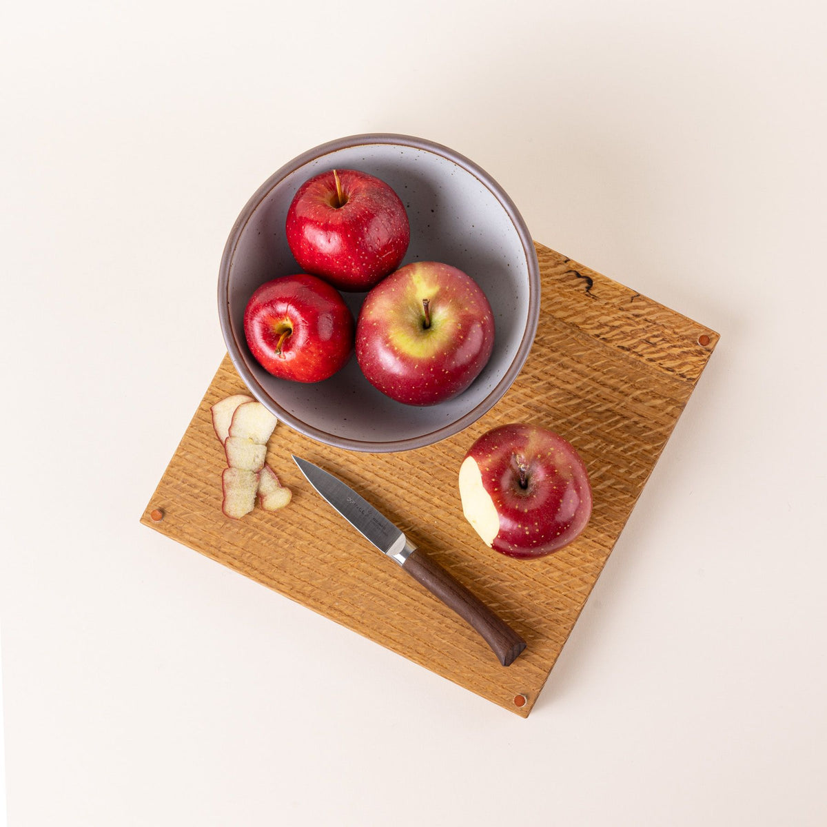 A cutting board and a bowl filled with apples with a small paring knife with a sharp steel blade and a beveled dark wood handle