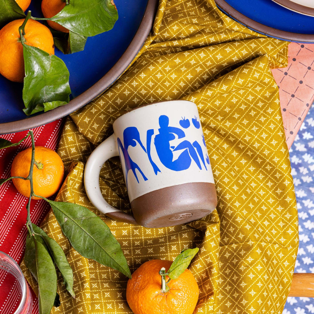 East Fork’s The Mug in a warm off white color with handle with a blue decal of Henri Matisse’s Femmes et singes, and a unglazed rim and bottom base, sits on a table with gold fabric and oranges.