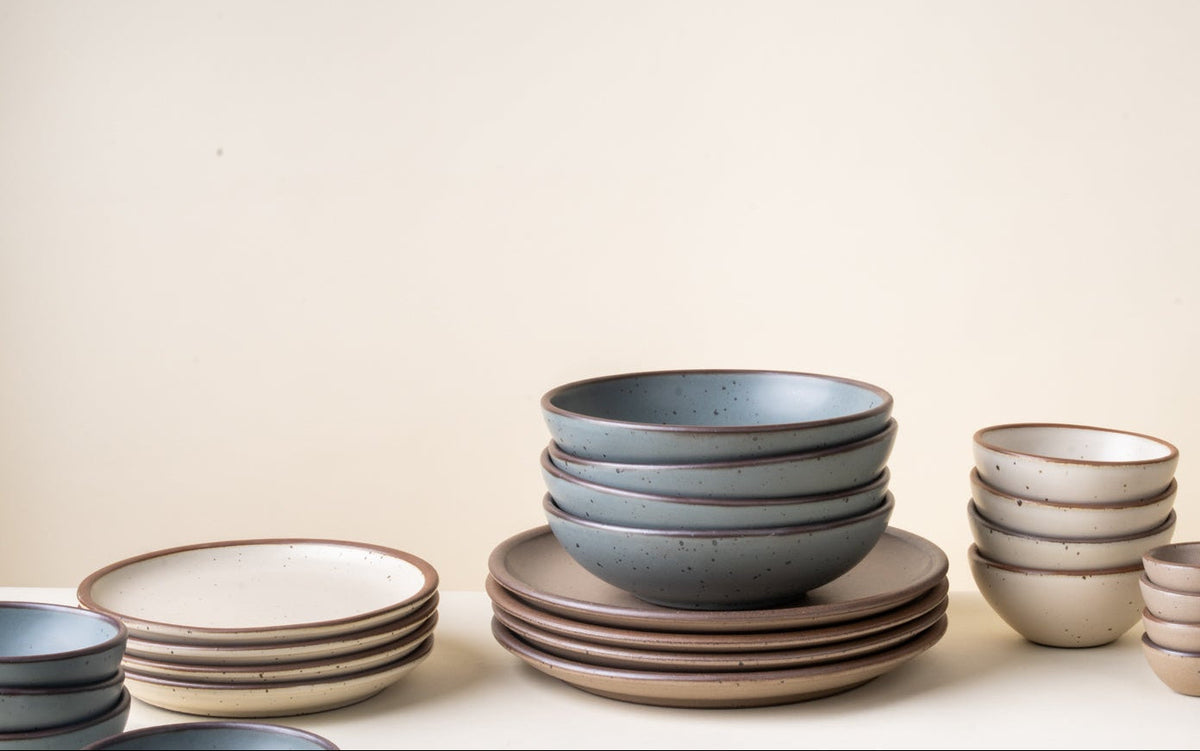 Stacks of ceramic pottery in blue-grey, off-white, and pale warm brown colors with iron speckles.