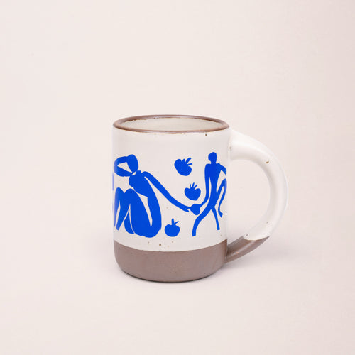 East Fork’s The Mug in a warm off white color with handle with a blue decal of Henri Matisse’s Femmes et singes, a bright blue, abstract cut-out figures of women sitting, monkeys, and pomegranate across the body of the mug