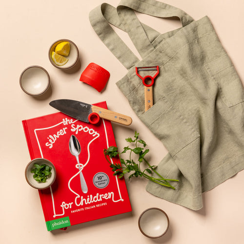 A styled flat-lay featuring a sage green linen apron with a red-handled vegetable peeler in its pocket, alongside a red children's recipe book titled 'The Silver Spoon for Children' by Phaidon, a small knife, herbs, and bowls on a light beige background.