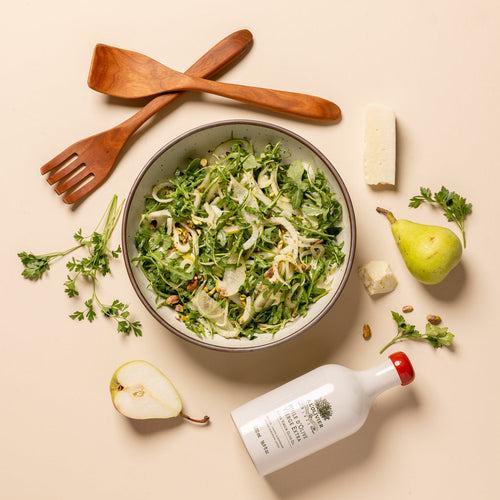 A styled flat-lay of a green salad in a ceramic bowl, surrounded by wooden serving utensils, a bottle of olive oil, pears, parmesan cheese, and parsley on a warm beige background.