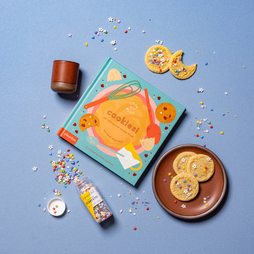 A styled flat-lay of a colorful book titled 'cookies!' on a light blue background, surrounded by cookies on a plate, sprinkles, a small cup in a terracotta, and a jar of sprinkles.