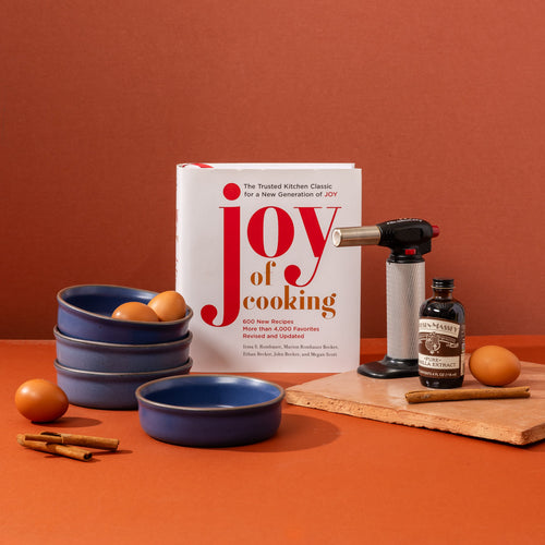 A cookbook titled "Joy of cooking" stands on a warm cream background alongside 4 blue colored, ceramic ramekins, a torch lighter, and a small bottle of vanilla, with eggs and cinnamon sticks against an orange background.