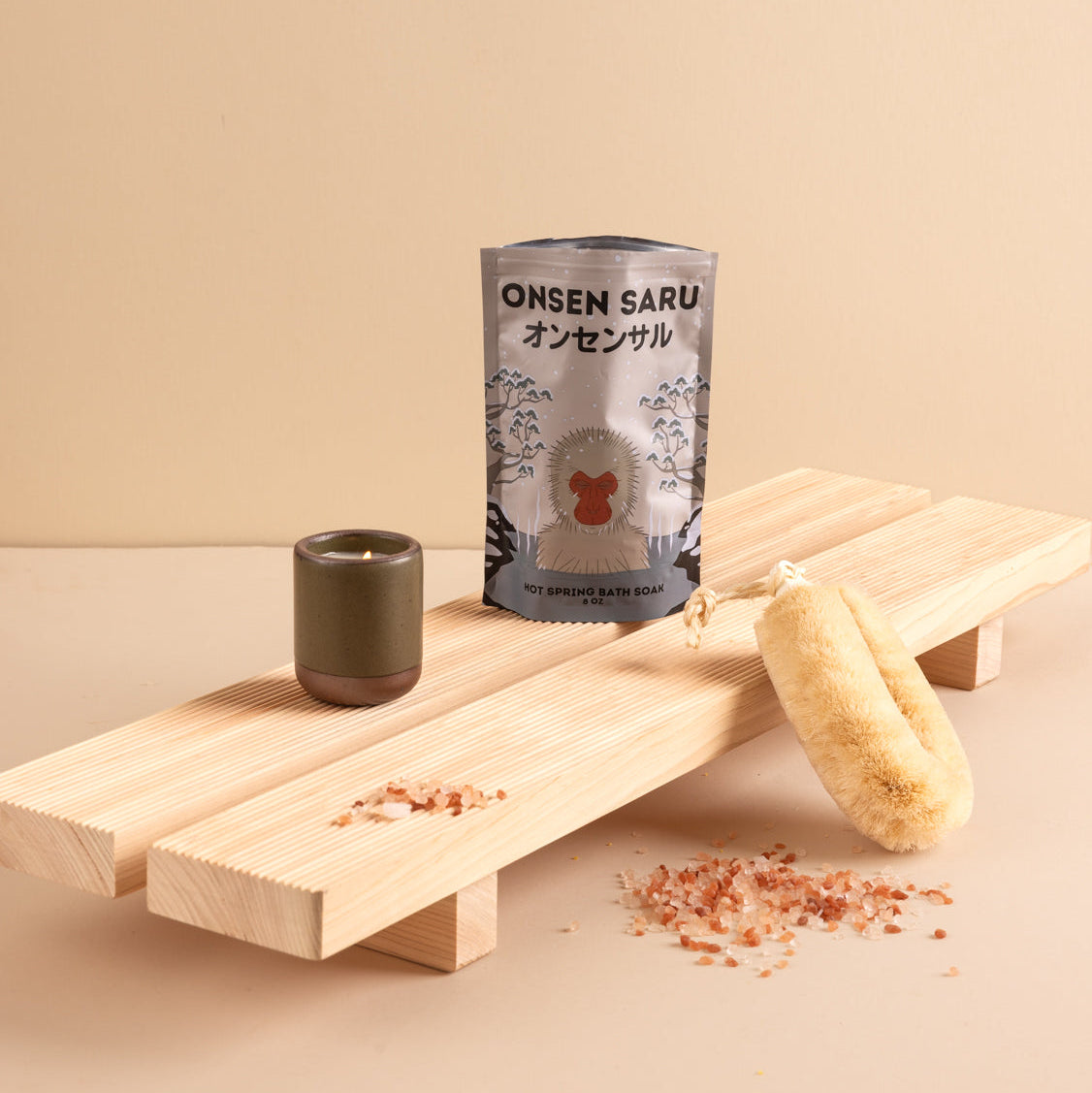 A packaged bath product labeled ONSEN SARU stands on a light wood platform beside a small dark green ceramic candle and a natural sponge, with scattered pink salt crystals on a beige background.