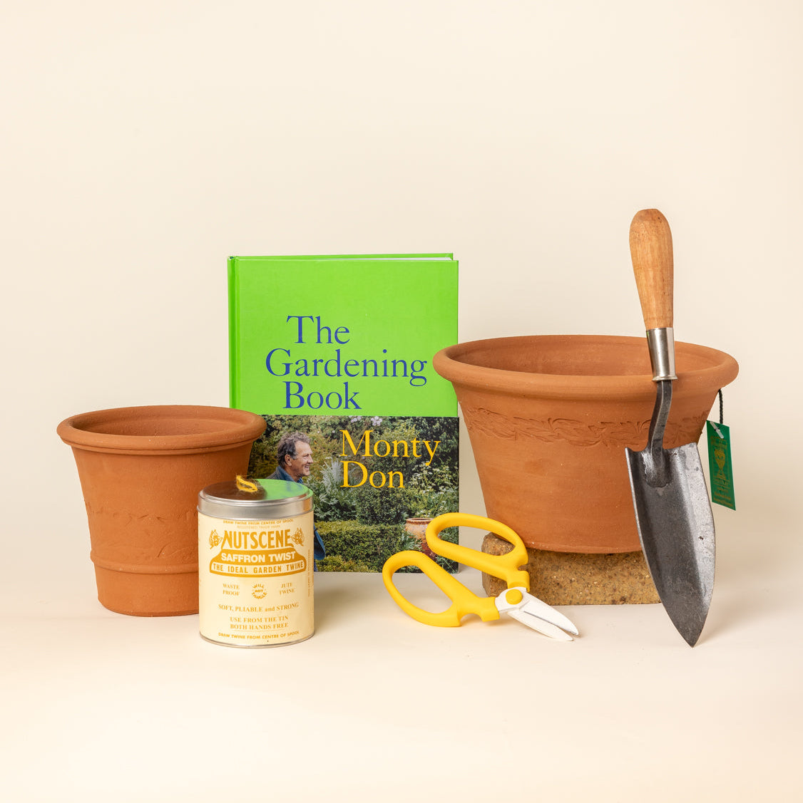 Gardening starter kit including two terracotta pots, a hand trowel, yellow-handled flower scissors, a tin of gardening twine and Monty Don's 'The Gardening Book' arranged on a light beige background.