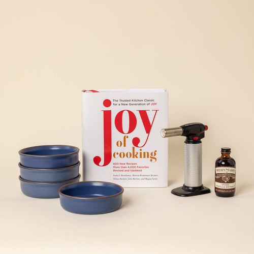 A cookbook titled "Joy of cooking" stands on a warm cream background alongside 4 grounded blue colored, ceramic ramekins, a torch lighter, and a small bottle of vanilla. 