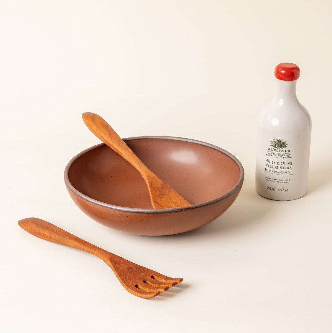 A large, shallow ceramic serving bowl in a matte terracotta color featuring an unglazed rim, paired with a bottle of olive oil and two wooden serving utensils, against a cream background.