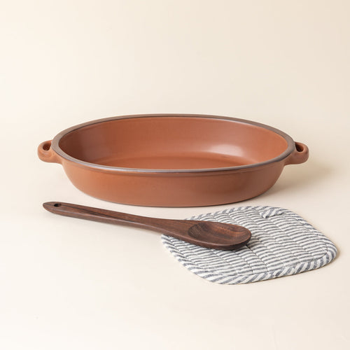 A large ceramic oval baker in a cool terracotta color with a wood spoon and striped pot holder.