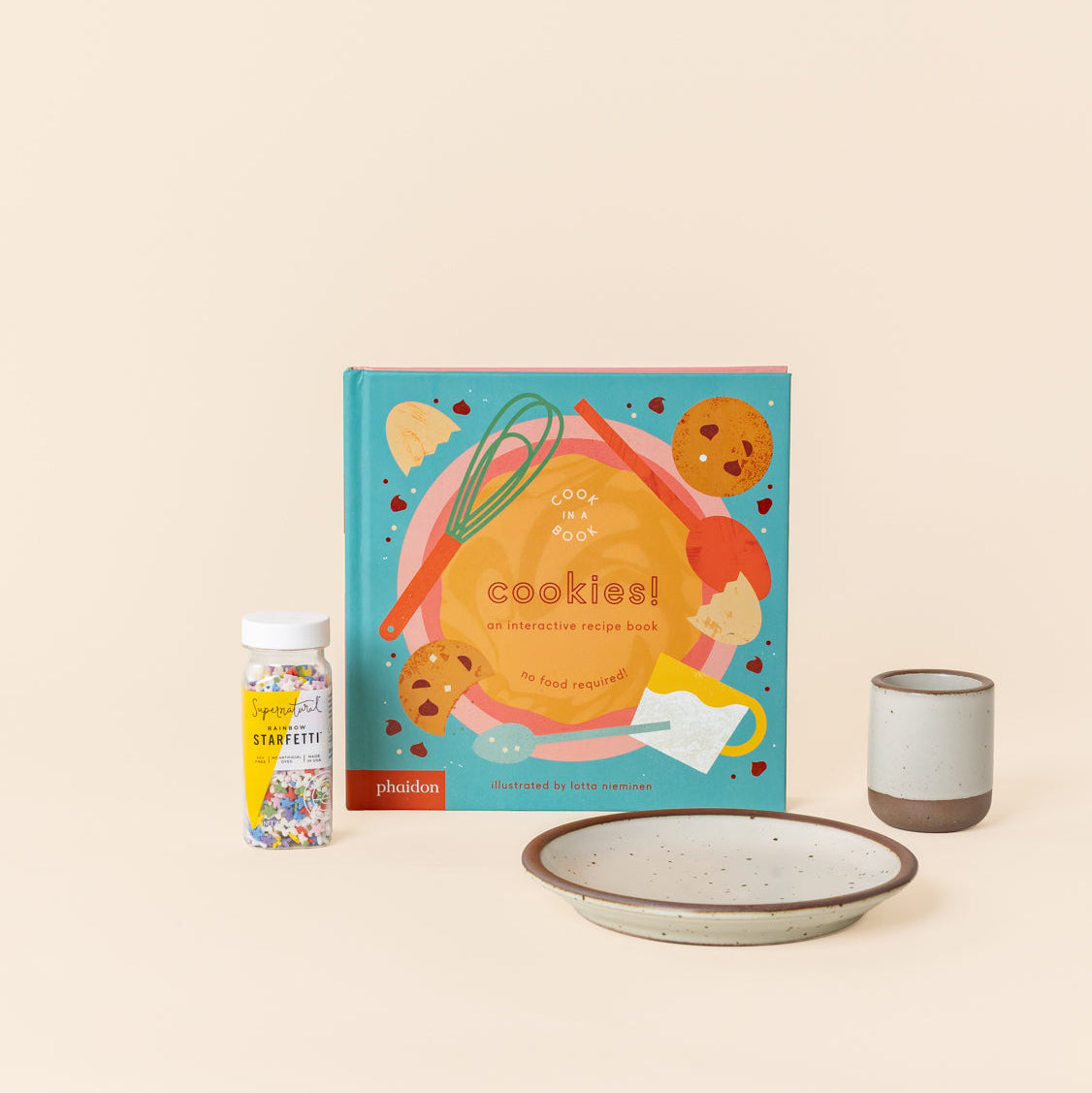A colorful book titled 'cookies!', a bottle of rainbow sprinkles, and a matching tiny cup and cake plate in a warm off-white color with unglazed rims.