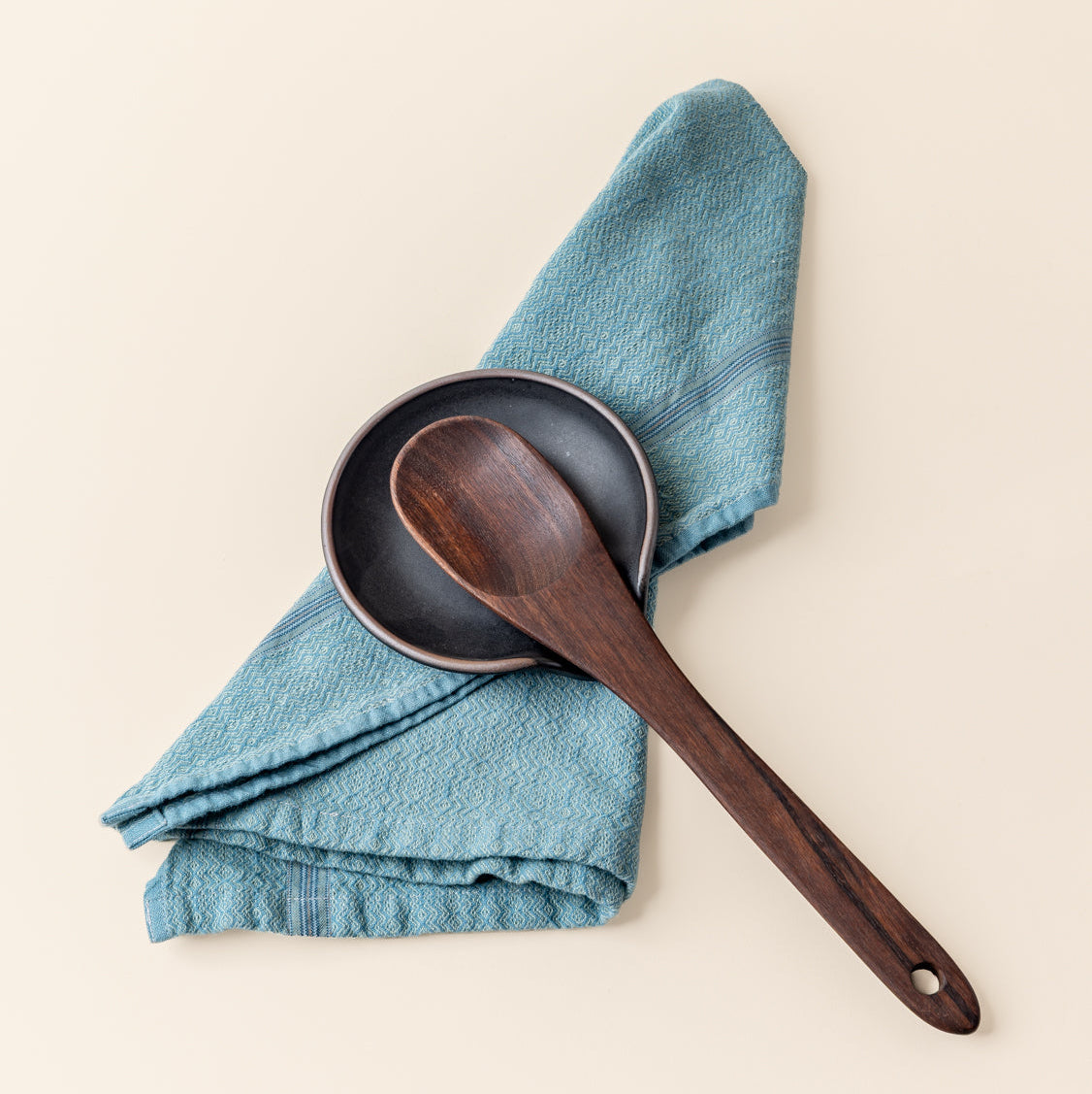 A ceramic spoon rest in a graphite black color with an unglazed rim, a blue kitchen towel, and a wooden kitchen spoon.