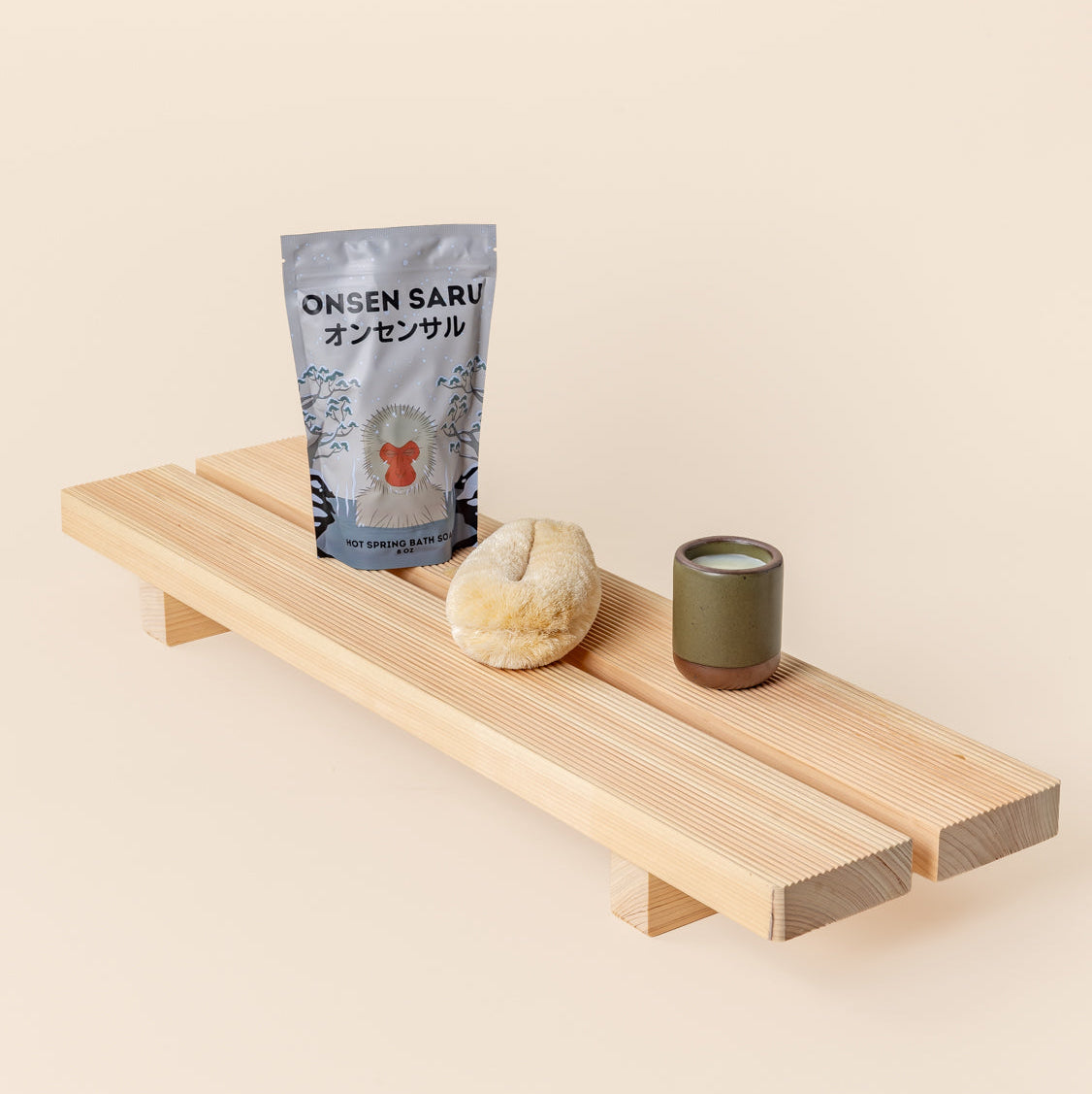 A silver bag of hot spring bath soak, a natural bath sponge, and a small olive green ceramic cup arranged on a light wooden bath tray against a cream background.