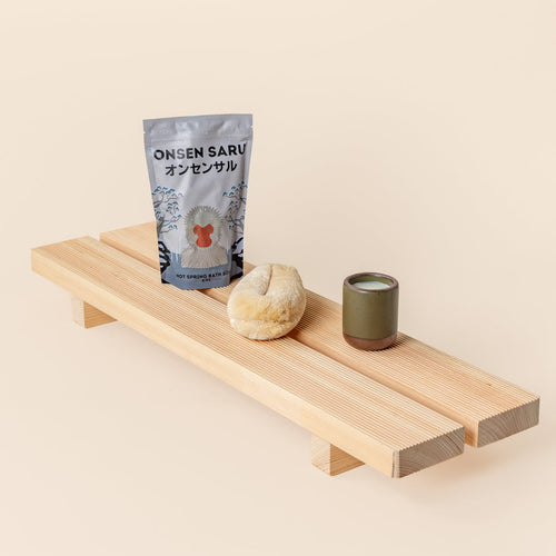 A silver bag of hot spring bath soak, a natural bath sponge, and a small olive green ceramic cup arranged on a light wooden bath tray against a cream background.