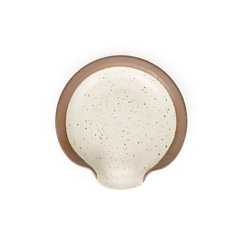 A circular ceramic spoon rest in a warm off-white color featuring iron speckles and unglazed ridge rim