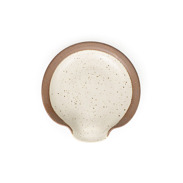 A circular ceramic spoon rest in a warm off-white color featuring iron speckles and unglazed ridge rim